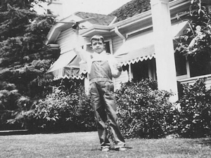 A young boy in a button up shirt and overalls standing in the front yard of his home, holding a toy plane in one hand high above his head.