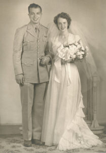 A young man in 1940s-era Army dress uniform holding hands with a young woman wearing a white wedding gown, bridal veil, and a large bouquet of white flowers. They are both smiling.