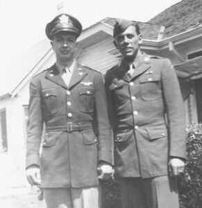 Two young men stand outside their childhood home together in military uniforms.