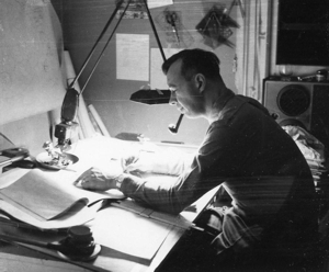 A man bent over an engineering table, examining a paper and surrounded by stacks of other documents.