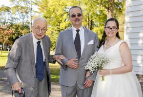 An elderly man and a middle-aged man stand in suits next to a young woman in a wedding gown.